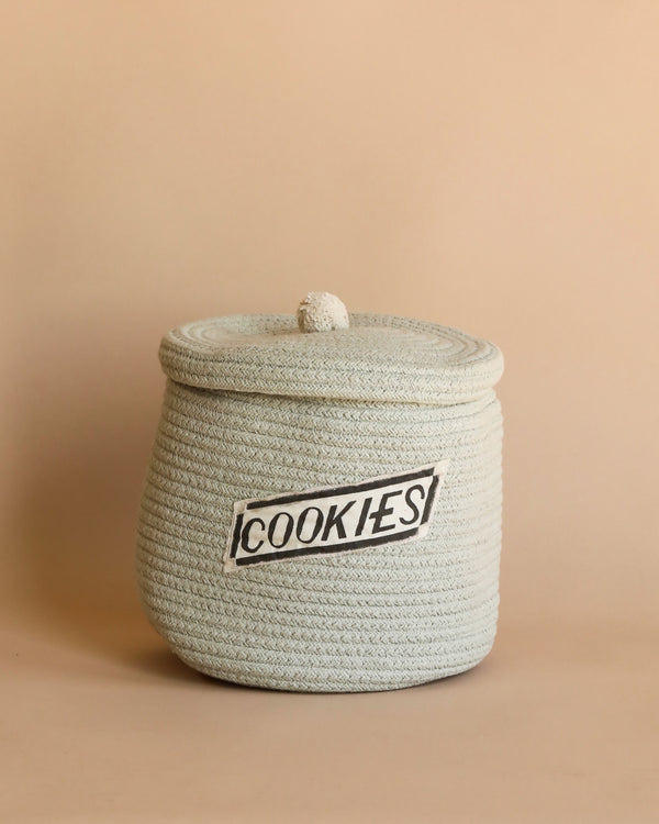 A round, handcrafted Basket cookie jar with a lid and a label that reads "cookies" in black text, set against a soft peach background.
