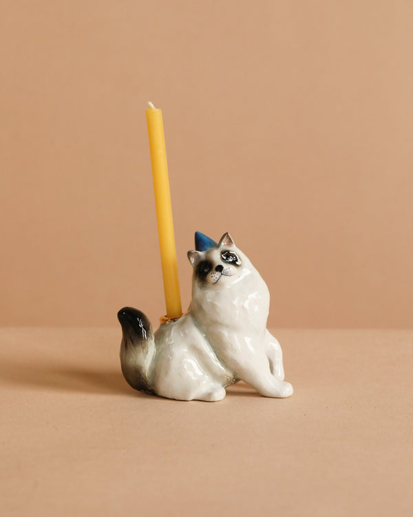 The Ragdoll Cat Cake Topper is a ceramic figurine with a blue party hat and a yellow candle on its back, displayed on a plain beige background—ideal for cat-themed parties.