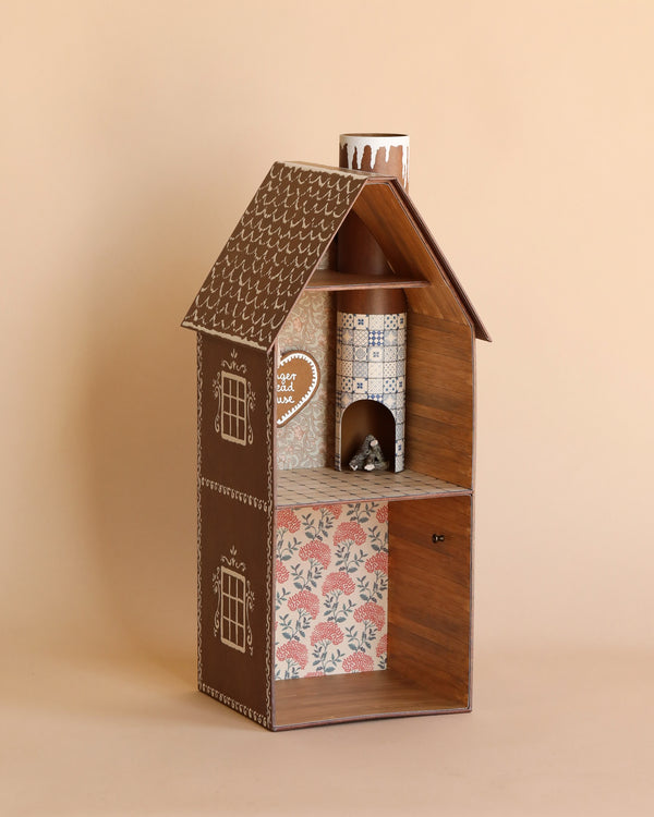 A decorative Maileg Cardboard Two-Story Gingerbread House with a quaint design, featuring a brown roof, textured walls, and floral wallpaper. The house has two visible floors with detailed windows and a small front porch.