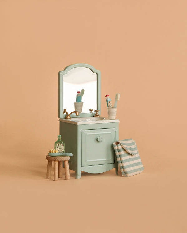 The Maileg Bathroom Set features a mint green sink dresser with mirror, two toothbrushes in a cup, toothpaste, soap, striped bag, and brush on a table and stool—all displayed against a plain beige background.