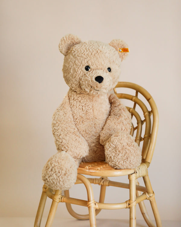 A plush beige Steiff, XL Jimmy Teddy Bear, 22 Inches sitting upright on a woven rattan chair with a light beige wall in the background. The teddy bear appears soft and has a friendly expression.