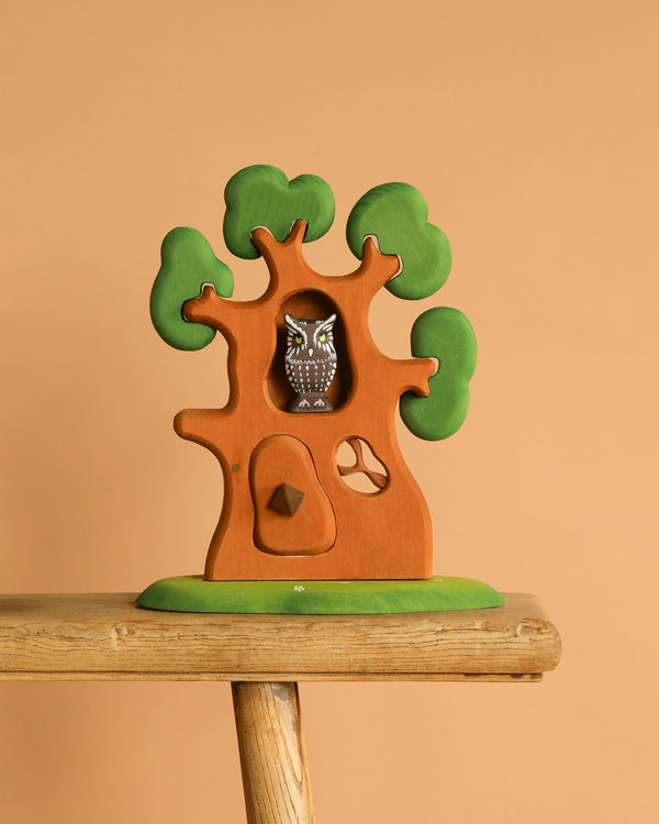 The Bumbu Handmade Wooden Ancient Oak Tree with Owl Set, featuring green leaves and an owl in a hollow, is displayed on a wooden stool against a beige background.