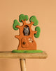 The Bumbu Handmade Wooden Ancient Oak Tree with Owl Set, featuring green leaves and an owl in a hollow, is displayed on a wooden stool against a beige background.
