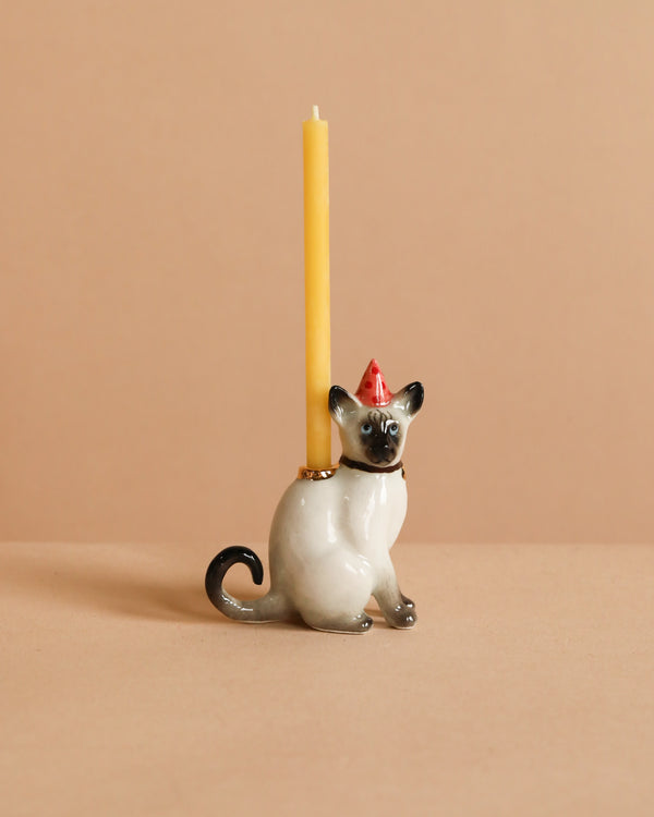 The Siamese Cat Cake Topper features a ceramic cat wearing a red party hat and holding a yellow taper candle on its back, ideal for cat-themed celebrations. The topper is displayed against a plain beige background.
