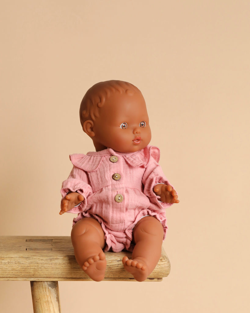 The Minikane Doll (13") - Leonie, made of phthalate-free vinyl with brown skin, wears a pink ruffled outfit with buttons. It sits on a wooden stool against a beige background, inviting imaginative playtime with its gently outstretched arms and legs.