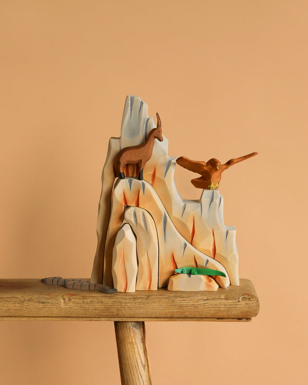 The Bumbu Handmade Wooden Mountain Cliff Scene features a goat atop the peak, an eagle with outstretched wings, and a bush at the base, all crafted from wood and displayed on a beige background over a wooden surface.