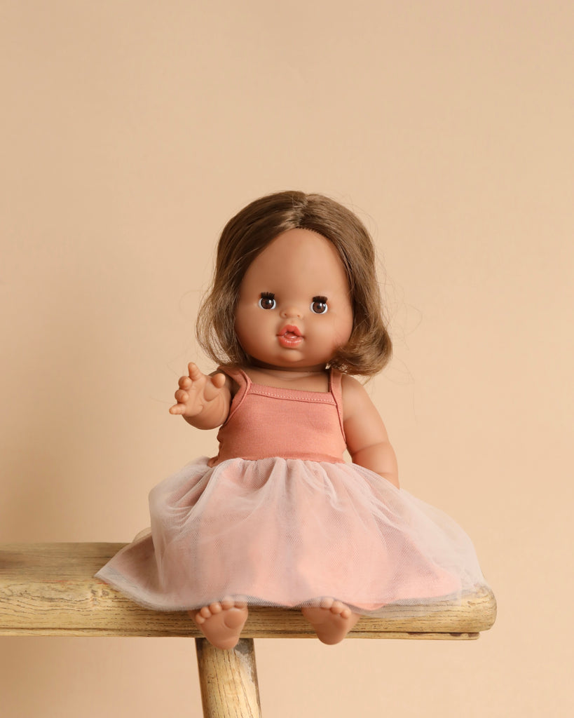 The Minikane Baby Doll (13") - Leopoldine with Clothing, featuring long brown hair and a vanilla scent, sits on a wooden stool in a pink dress with a tulle skirt against a beige background. Its arm is raised as if waving.