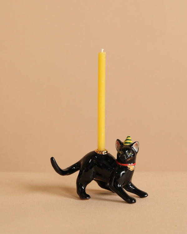 The Black Cat Cake Topper is a black cat-shaped candle holder with a yellow candle on its back, wearing a striped party hat and red collar, ideal for cake decoration and set against a plain beige background.