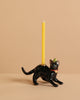 The Black Cat Cake Topper is a black cat-shaped candle holder with a yellow candle on its back, wearing a striped party hat and red collar, ideal for cake decoration and set against a plain beige background.