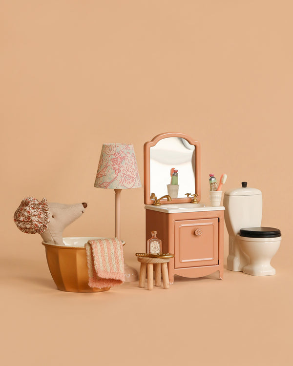A Maileg Bathroom Set is arranged on a beige background, featuring a Wellness Mouse in a gold bathtub with pink towel, sink dresser with mirror, lamp, toiletries on a stool, and a white toilet.
