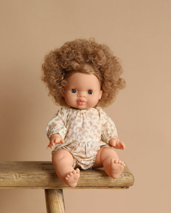 The Minikane Baby Doll (13"), Anais, with curly brown hair and an anatomically correct design, sits on a wooden bench against a beige background. Dressed in a patterned off-white romper, the doll displays a neutral expression.