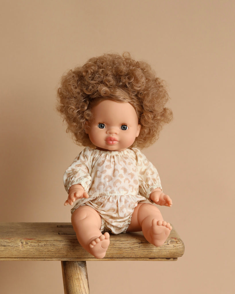 The Minikane Baby Doll (13"), Anais, with curly brown hair and an anatomically correct design, sits on a wooden bench against a beige background. Dressed in a patterned off-white romper, the doll displays a neutral expression.