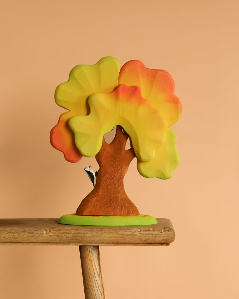 The Bumbu Handmade Wooden Large Oak Autumn and Woodpecker Set features a colorful hand-painted oak with yellow, orange, and green leaves, plus a small black and white woodpecker perched on the trunk, against a solid light peach background.