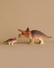 Two figurines from the Bumbu Handmade Wooden Prehistoric Set, one large and one small triceratops, painted in brown, pink, and white, face each other on a plain beige background—ideal for educational and imaginative play.