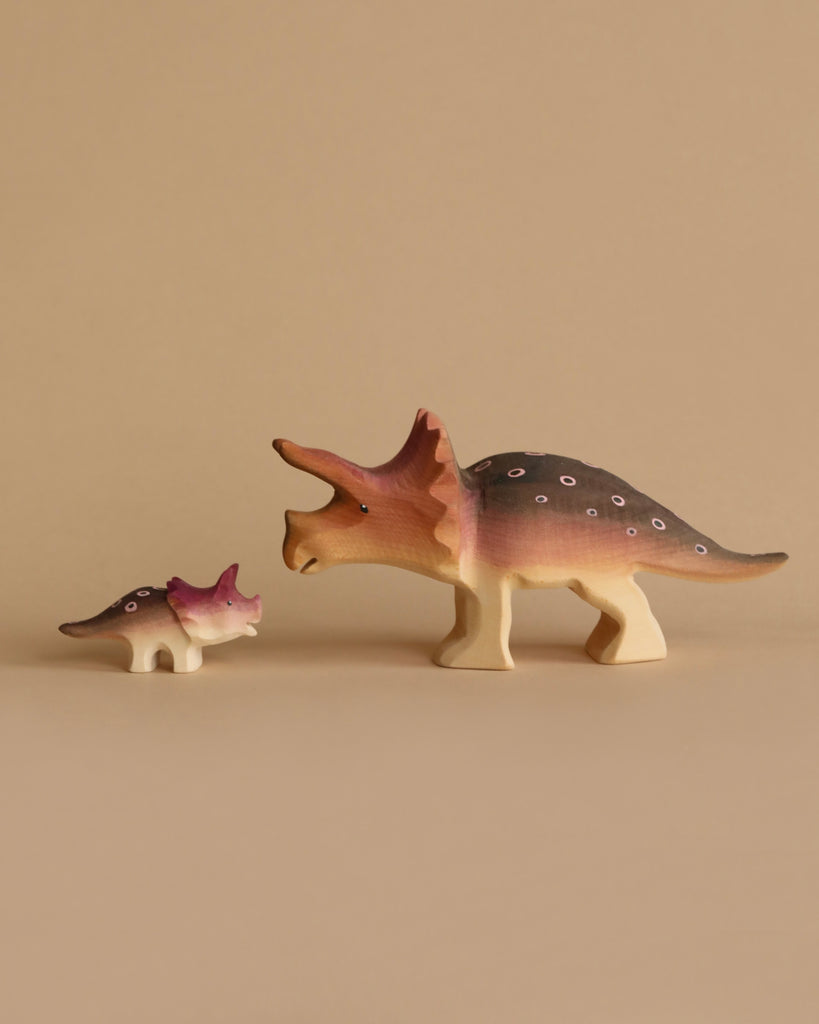 Two figurines from the Bumbu Handmade Wooden Prehistoric Set, one large and one small triceratops, painted in brown, pink, and white, face each other on a plain beige background—ideal for educational and imaginative play.