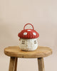 A whimsical Olli Ella | Red Mushroom Basket designed to resemble a mushroom, with a red cap and white base, resting on a rustic wooden stool against a plain beige background.