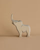 A light wooden toy elephant with its trunk raised stands against a beige background. Handcrafted using sustainable materials, this charming piece is reminiscent of the Ostheimer Small Elephant - Trumpeting, featuring simplicity and minimal details while casting a gentle shadow on the surface.