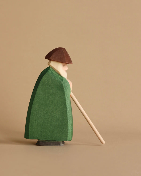 A handcrafted wood figurine of a bearded man wearing a brown hat, green cloak, and holding a wooden staff. The Ostheimer Shepherd With Staff is positioned sideways against a plain beige background, embodying the charm of sustainable materials.