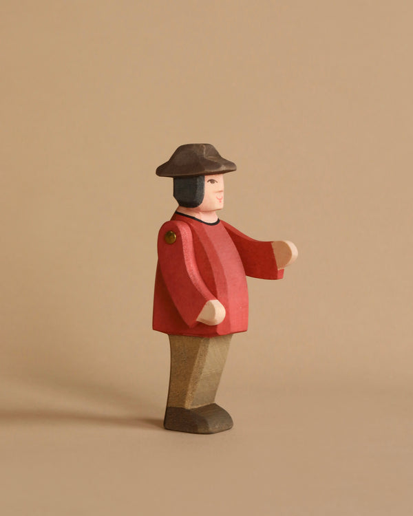 An Ostheimer Farmer from the Ostheimer brand stands against a neutral beige background. Dressed in a red top, brown pants, and a black hat, its arms extend outward slightly. With minimalistic facial details and a simple, handcrafted appearance, it invites imaginative play.