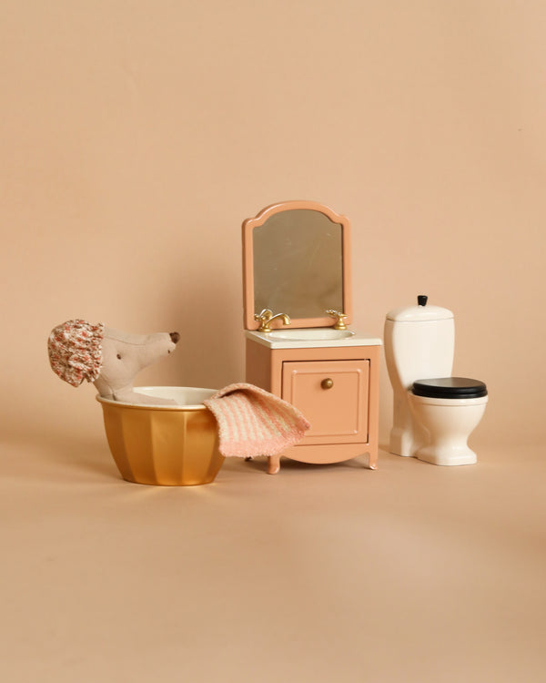 Miniature bathroom setup with a pink vanity, a gold-colored bathtub, a white toilet, and a Maileg Bathroom Starter Set mouse sitting on the tub, all against a beige background.