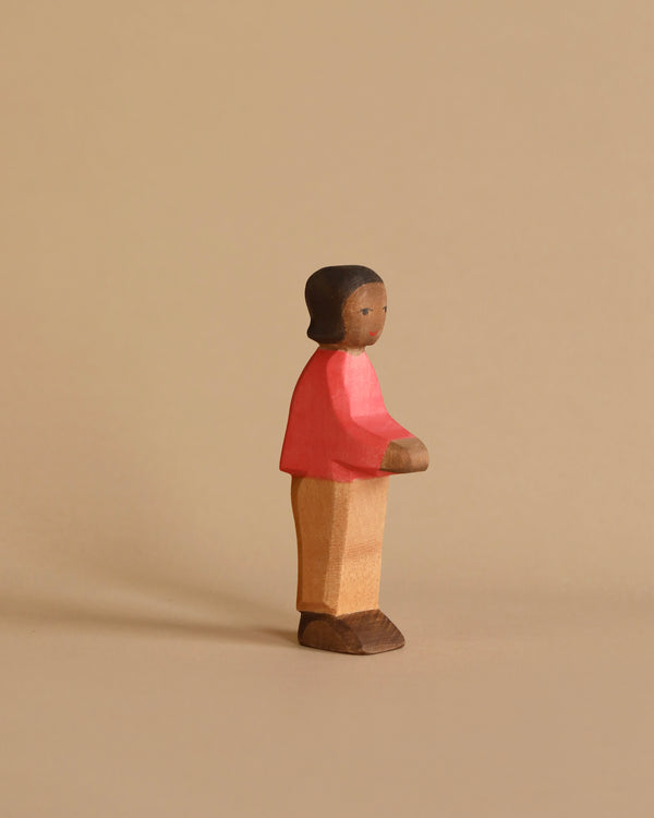 A small, handcrafted wooden figurine of a person stands against a beige background. The figure, dressed in a red sweater, tan pants, and brown shoes, has a simple, minimalist design with a smooth texture and neutral facial expression. Made using sustainably sourced materials, it evokes the charm of an Ostheimer Son, Red Sweater.