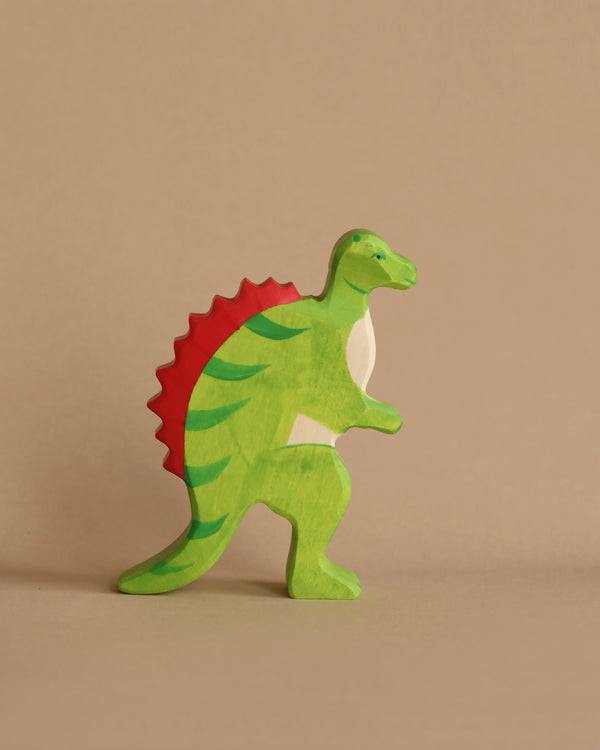 A colorful, handcrafted wood dinosaur toy with light green body, red spines along its back, and white underbelly. This charming Holztiger Spinosaurus Dinosaur stands upright against a plain beige background, showcasing the fine craftsmanship made in Europe.