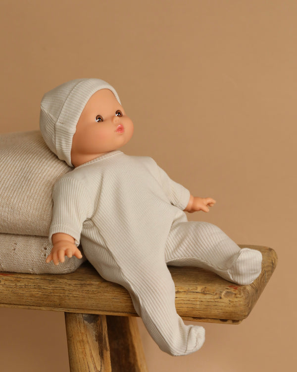 A Minikane Soft Body Doll (11") - Alice With Clothing sits on a wooden bench in a white outfit and hat, against a neutral beige background. A textured cushion promises safe vinyl fun for imaginative playtime.