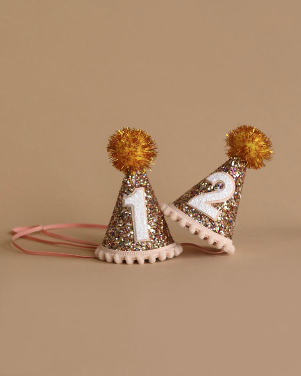 Two small, Confetti Glitter Party Hats with gold pom-poms and white numbers "1" and "2" on them are perfect for birthday celebrations. The hats, complete with pink elastic bands, are set against a plain beige background—ideal for festive photo shoots.