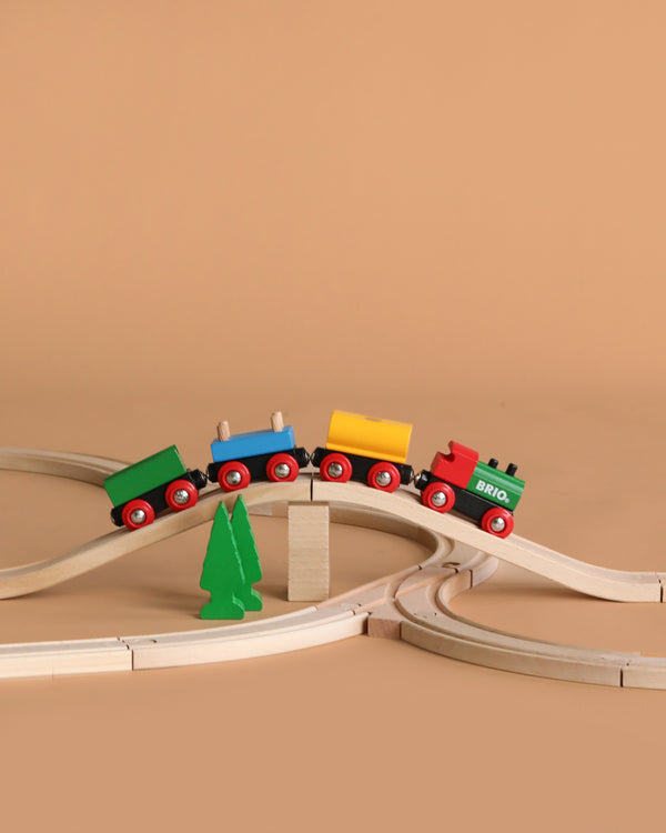 The BRIO World 65th Anniversary Train Set features a colorful wooden train with four cars on a raised curved track and a green tree-shaped piece, evoking classic playtime nostalgia against a simple light brown background.