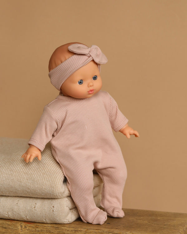 The Minikane Soft Body Doll (11") - Claire is dressed in a beige ribbed outfit with a matching bow headband and sits upright on two folded towels against a soft brown background, highlighting its charm and phthalate-free vinyl construction.