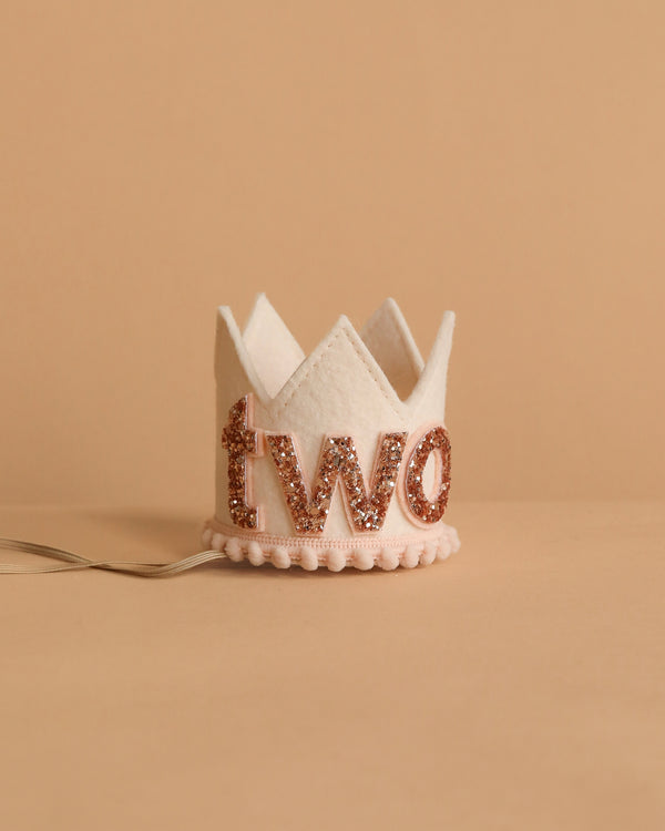 The Linen and Rose Crown is a small white felt crown with "two" in rose gold glitter, ideal for birthdays. It rests on a neutral beige surface and features an attached ribbon for easy wearing.