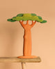 The Bumbu Handmade Wooden Extra Large Baobab Tree, featuring a thick brown trunk and layered green foliage, sits on a wooden surface against a beige background.