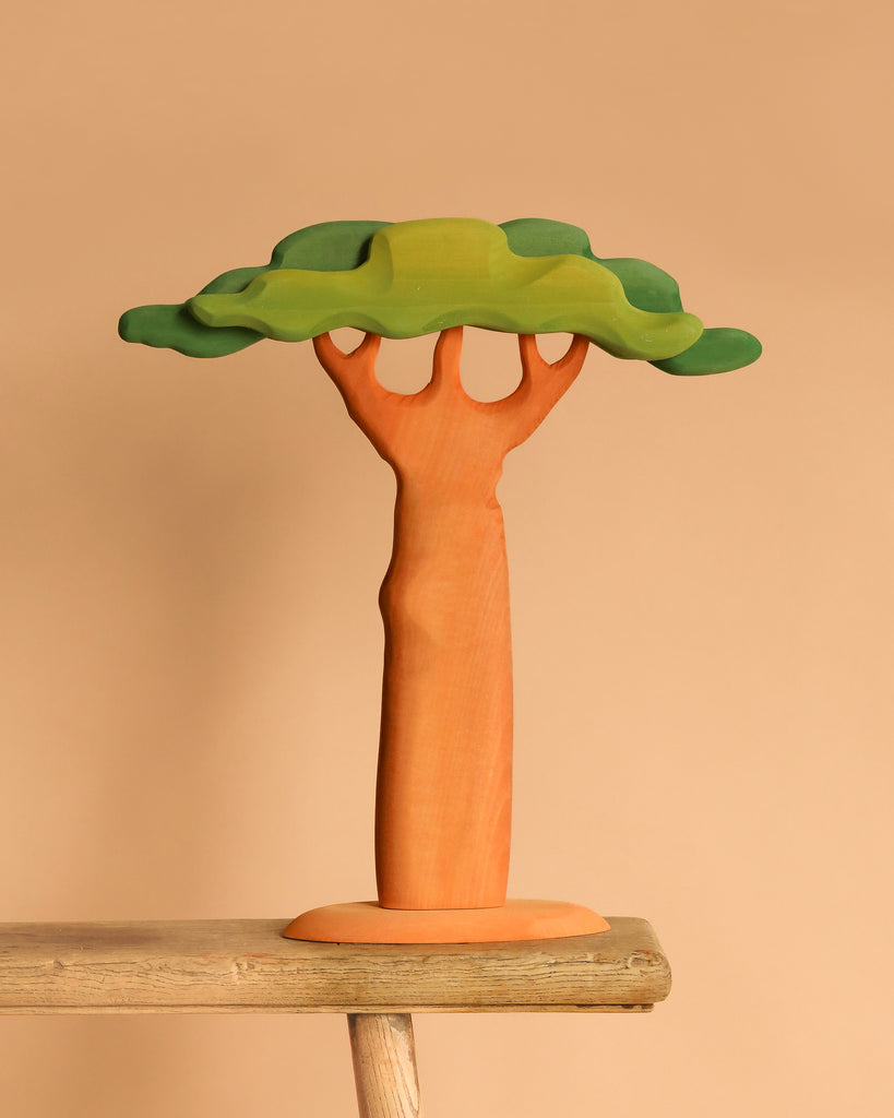 The Bumbu Handmade Wooden Extra Large Baobab Tree, featuring a thick brown trunk and layered green foliage, sits on a wooden surface against a beige background.