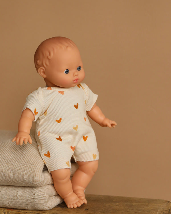 The Minikane Soft Body Doll (11") - Claire With Clothing, made from phthalate-free vinyl, has blue eyes and a soft body. The doll wears a cream onesie with orange hearts and is seated on a beige blanket against a plain brown background.