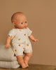 The Minikane Soft Body Doll (11") - Claire With Clothing, made from phthalate-free vinyl, has blue eyes and a soft body. The doll wears a cream onesie with orange hearts and is seated on a beige blanket against a plain brown background.