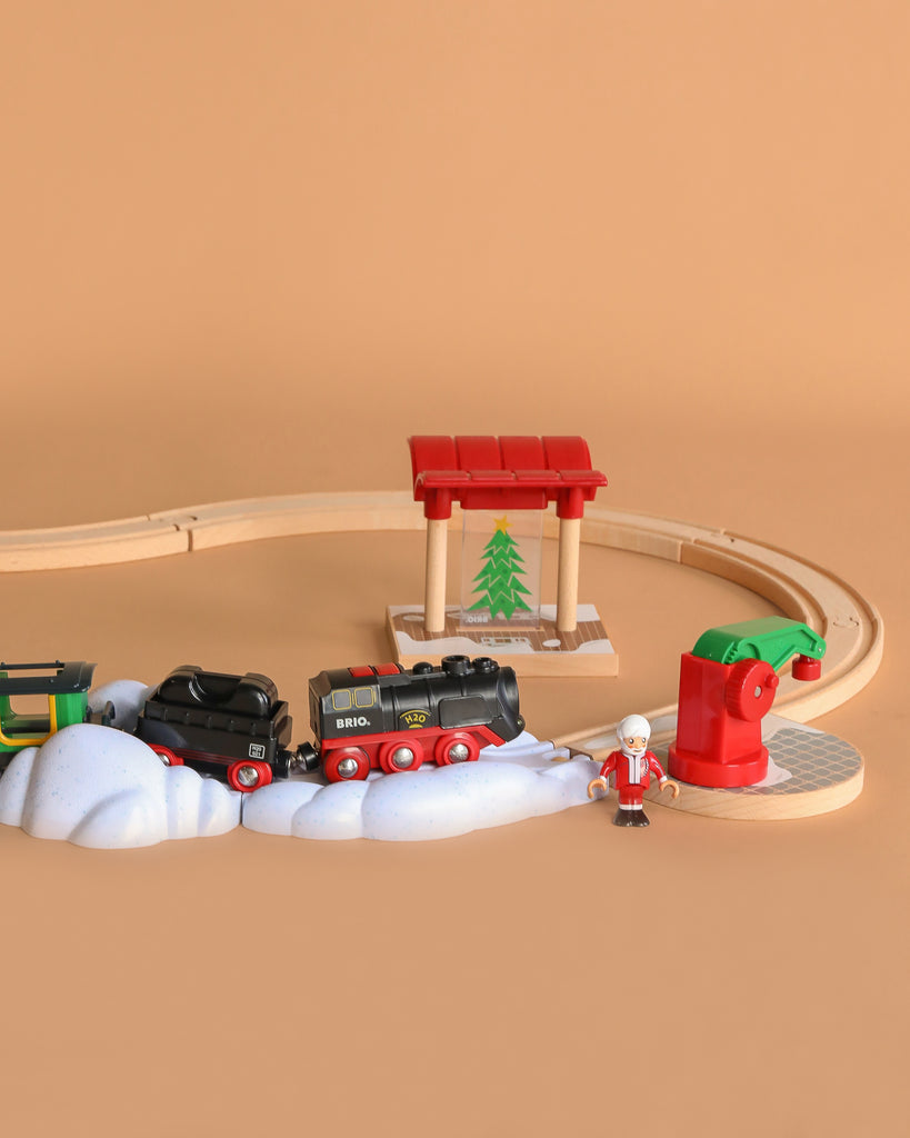 The BRIO World Christmas Steaming Train Set includes tracks, a black and red train, red station, snow decorations, green tree, red mailbox, and Santa figurine on a light brown background.