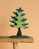 The Bumbu Handmade Wooden Bear and Forest Trees Set features a green, stylized tree with wavy branches, crafted from sustainably sourced wood, displayed on a rustic surface against a beige background.