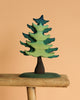 The Bumbu Handmade Wooden Prehistoric Set features a stylized green and blue tree sculpture with a dark trunk, crafted with non-toxic paint. Perfect for imaginative play, it sits beautifully on any wooden ledge against a neutral background.