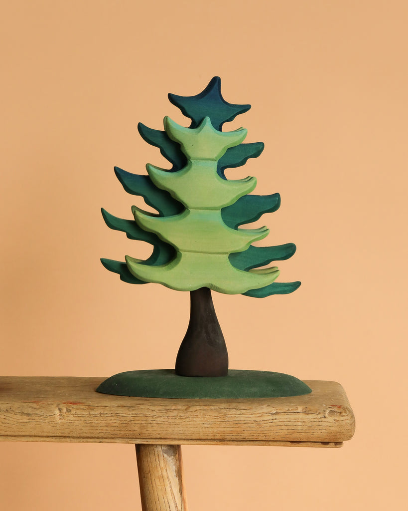 The Bumbu Handmade Wooden Prehistoric Set features a stylized green and blue tree sculpture with a dark trunk, crafted with non-toxic paint. Perfect for imaginative play, it sits beautifully on any wooden ledge against a neutral background.