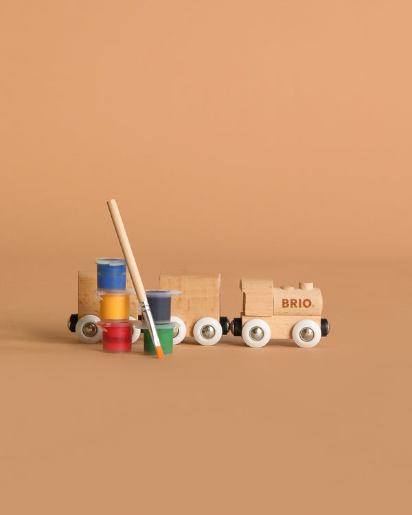 The BRIO World Paint Train with white wheels sits beside blue, yellow, red, and green paint pots and a paintbrush, all arranged on a plain beige background.