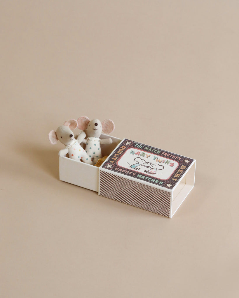 A charming scene showing two Maileg | Twin Baby Mice In Matchbox, representing newborn twins, peeking out of an open matchbox-style toy packaging. The packaging is adorned with playful illustrations and text.