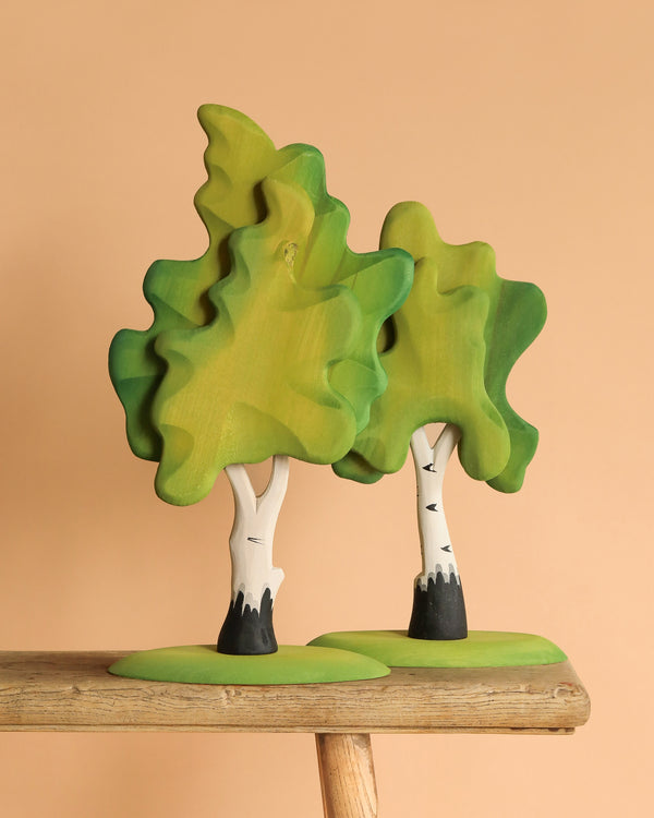 Two handmade toys from the Bumbu Handmade Wooden Extra Large Birch Tree Set, with white trunks and green canopies, stand on a wooden platform. Their stylized design features painted leaves and bases on a peach background.