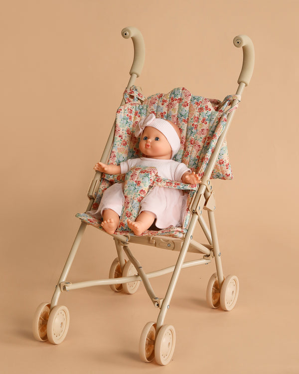 The Minikane Soft Body Doll (11") - Claire with Clothing and Stroller is shown wearing pink clothes and a hat, seated in a floral-patterned stroller with beige wheels and handles against a plain beige background.