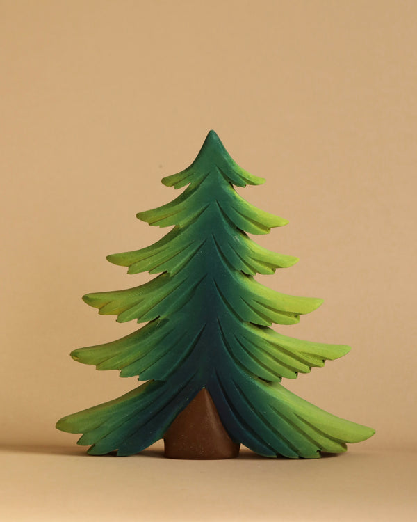 A Bumbu Large Wooden Fir Tree with multiple layered branches in varying shades of green stands against a neutral beige background. Handmade in Romania, the tree has a simple, smooth texture and a small, visible brown trunk at the bottom.