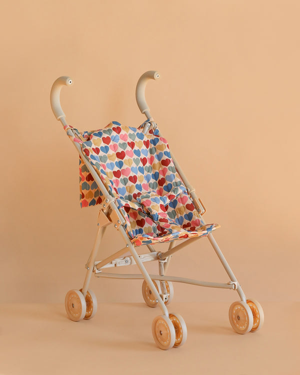 The Konges Sloejd Doll Stroller - Heart Kisses features a beige frame, double wheels, and a colorful heart-patterned seat, shown against a plain peach background.