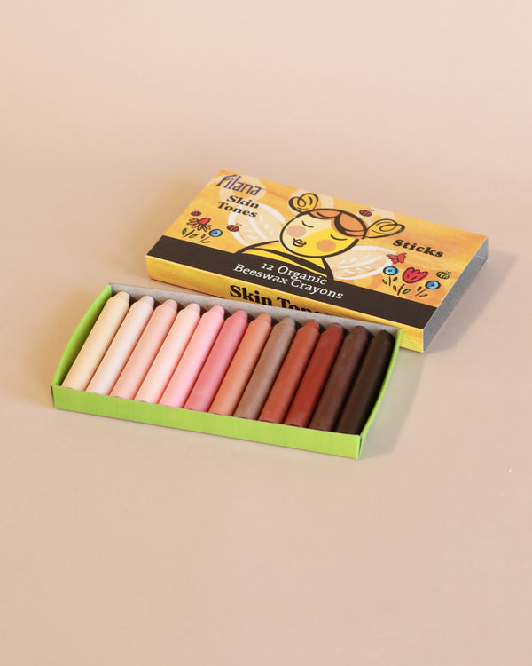 A box of Organic Beeswax Crayons: 12 Skin Tones in Sticks, containing organic beeswax crayons in various skin tones, displayed on a pale pink background.