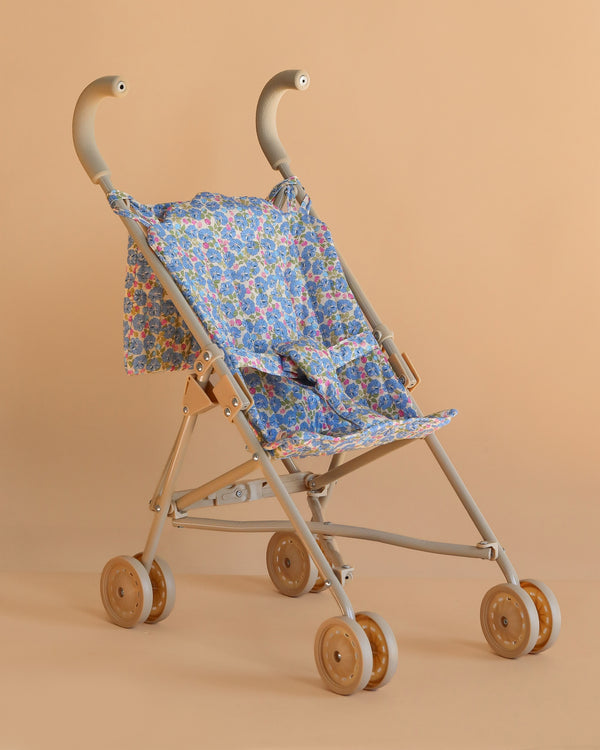 The Konges Sloejd Doll Stroller - Rose Flower features a beige frame, wooden double wheels, and a blue rose floral-patterned seat and backrest, shown against a plain peach background.