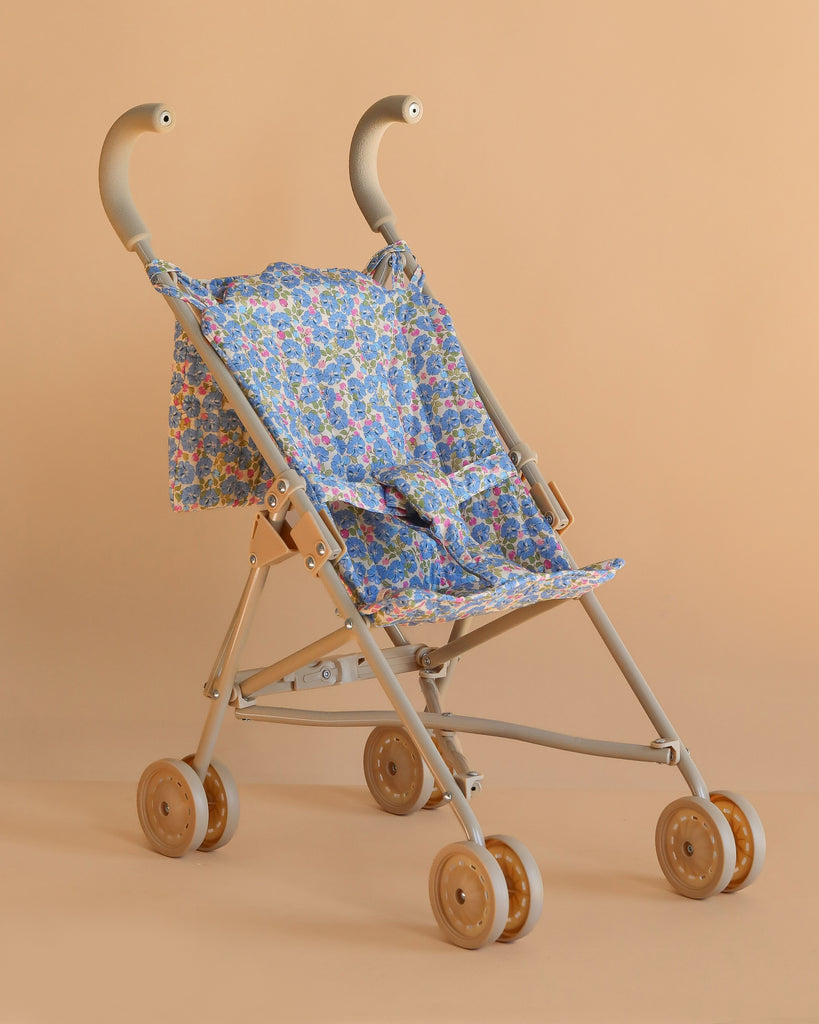 The Konges Sloejd Doll Stroller - Rose Flower features a beige frame, wooden double wheels, and a blue rose floral-patterned seat and backrest, shown against a plain peach background.