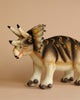 A Cream Triceratops Dinosaur Stuffed Animal stands against a beige background. Crafted by HANSA animals, it features realistic details with yellow and orange spots along its back, and a white underbelly and legs. The hand-sewn plush boasts three distinctive horns and a frilled neck, adding to its charm.