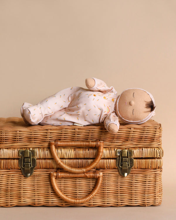 A Olli Ella x Odin Parker Dozy Dinkum - Bugsy Hopscotch in a 100% Organic Cotton white outfit with yellow patterns, lying on top of a woven wicker basket with closed lids and brass clasps, against a neutral background.